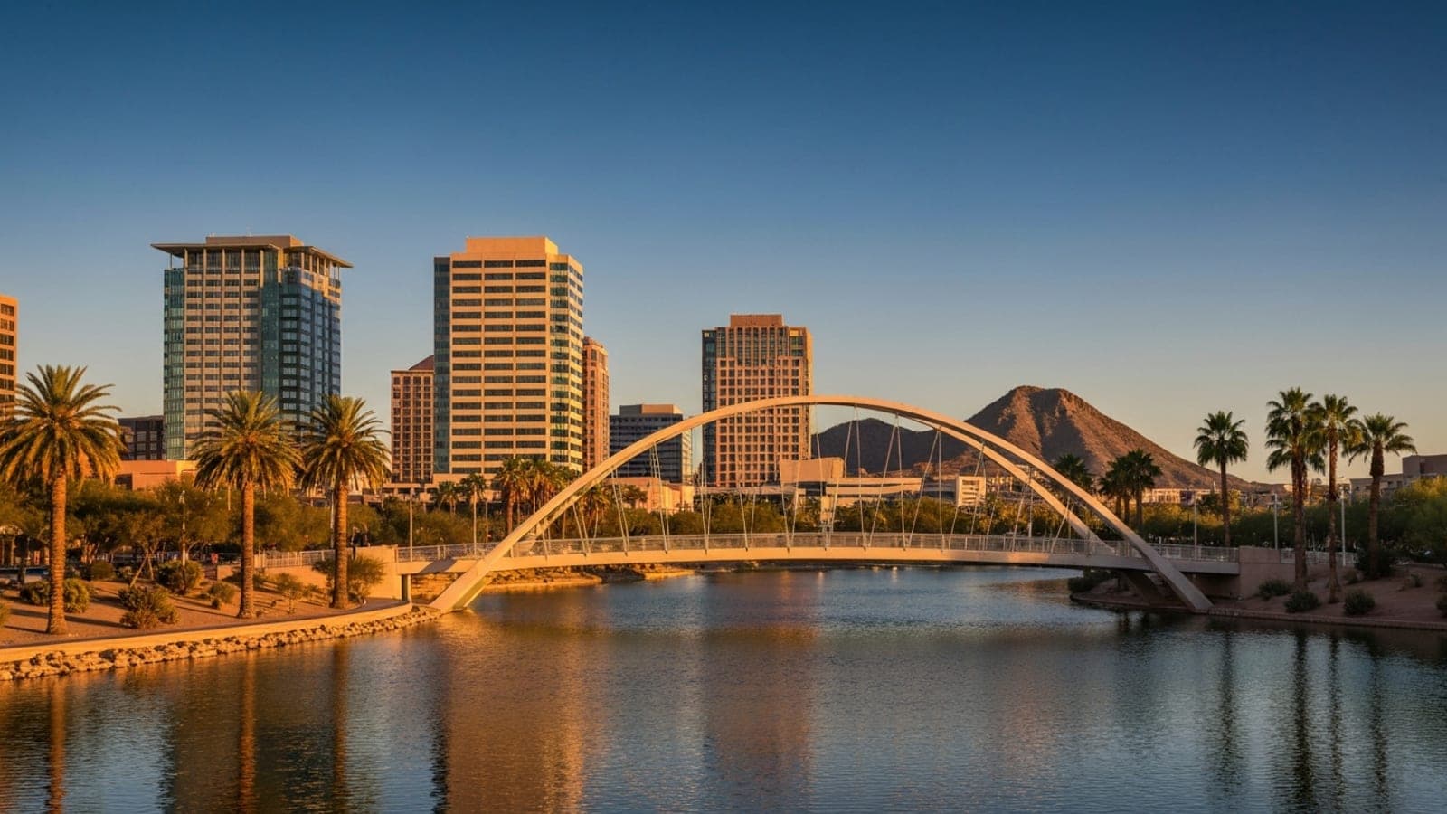 Editorial photograph of Tempe Town Lake at golden hour with the iconic pedestrian arch bridge, modern mid-rise downtown Tempe buildings in the background, and palm trees along the lakeshore.