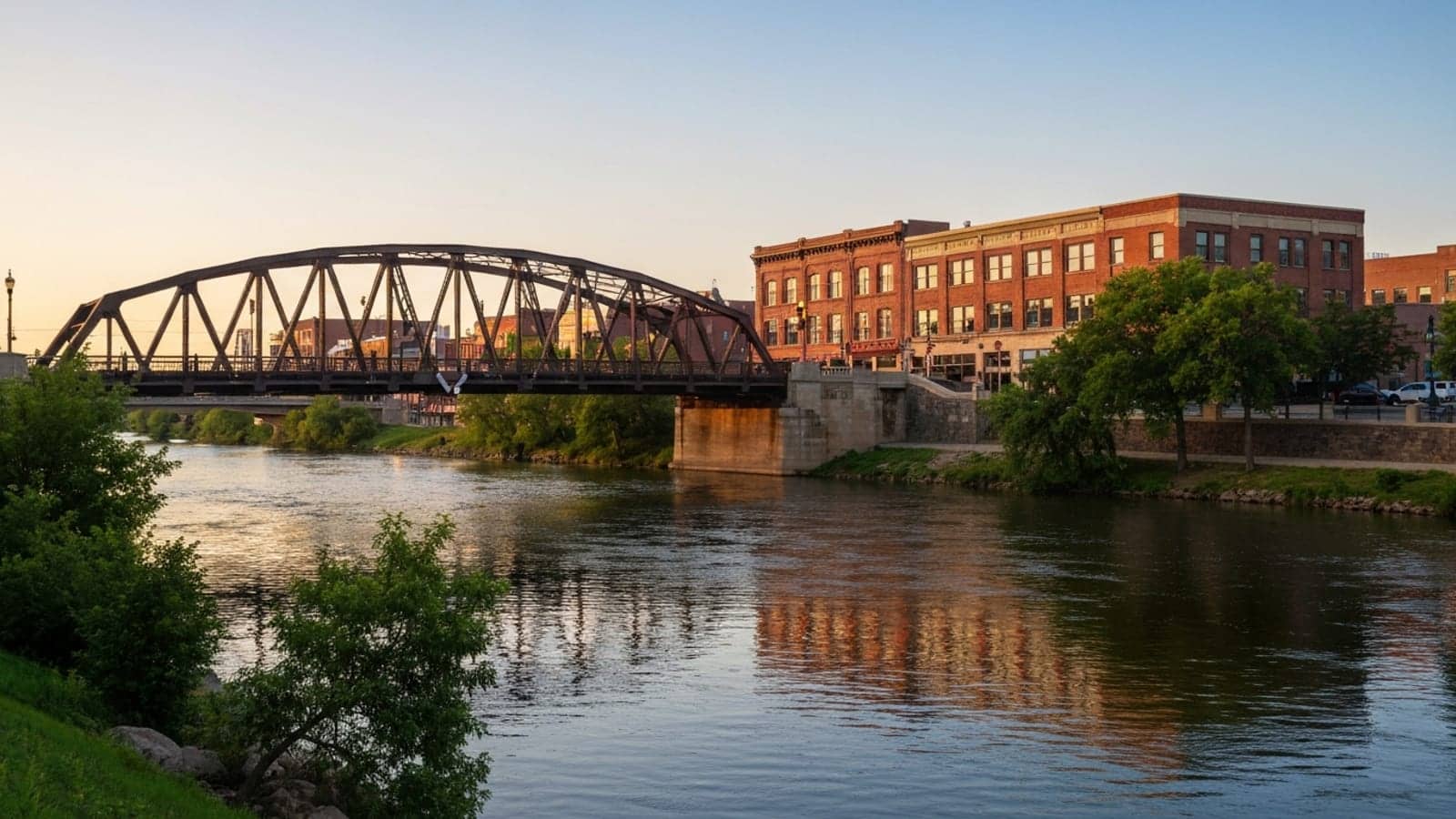 Editorial photograph of the Fox River bridges in downtown Elgin Illinois at golden hour with the historic Kimball Street truss bridge and brick downtown commercial buildings.